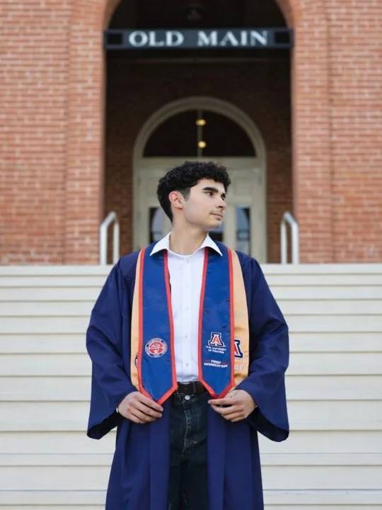 Nathanael Kenzler on the steps of Old Main at the University of Arizona, wearing a graduation gown and a first generation honor stole.